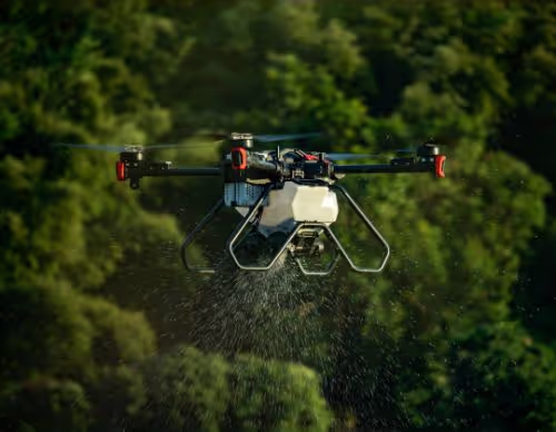 Agricultural drone spraying liquid over crops with green trees in background.